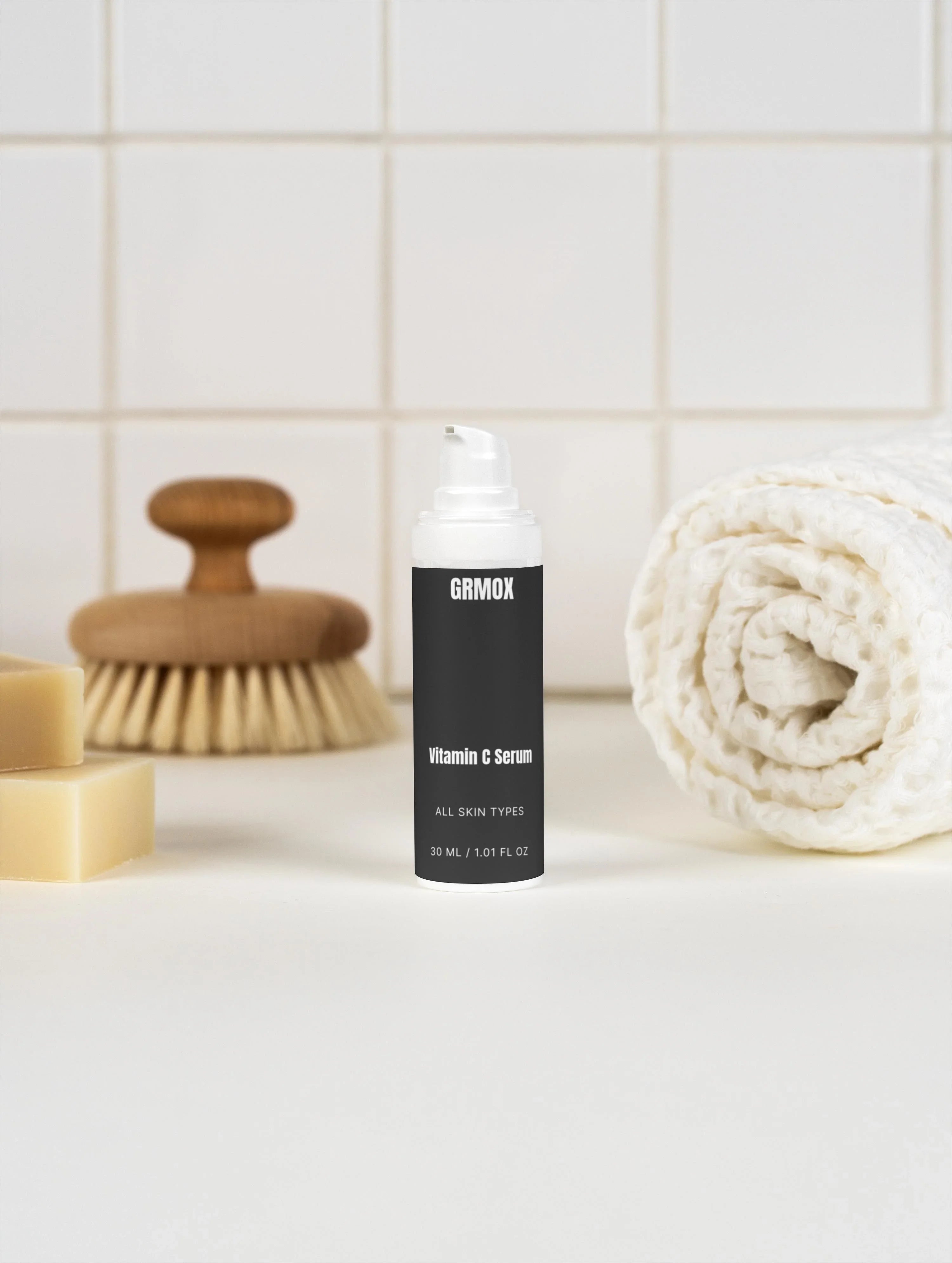GRMOX Vitamin C Serum for all skin types, 30ml bottle with pump, beside towel and brush on white tile background
