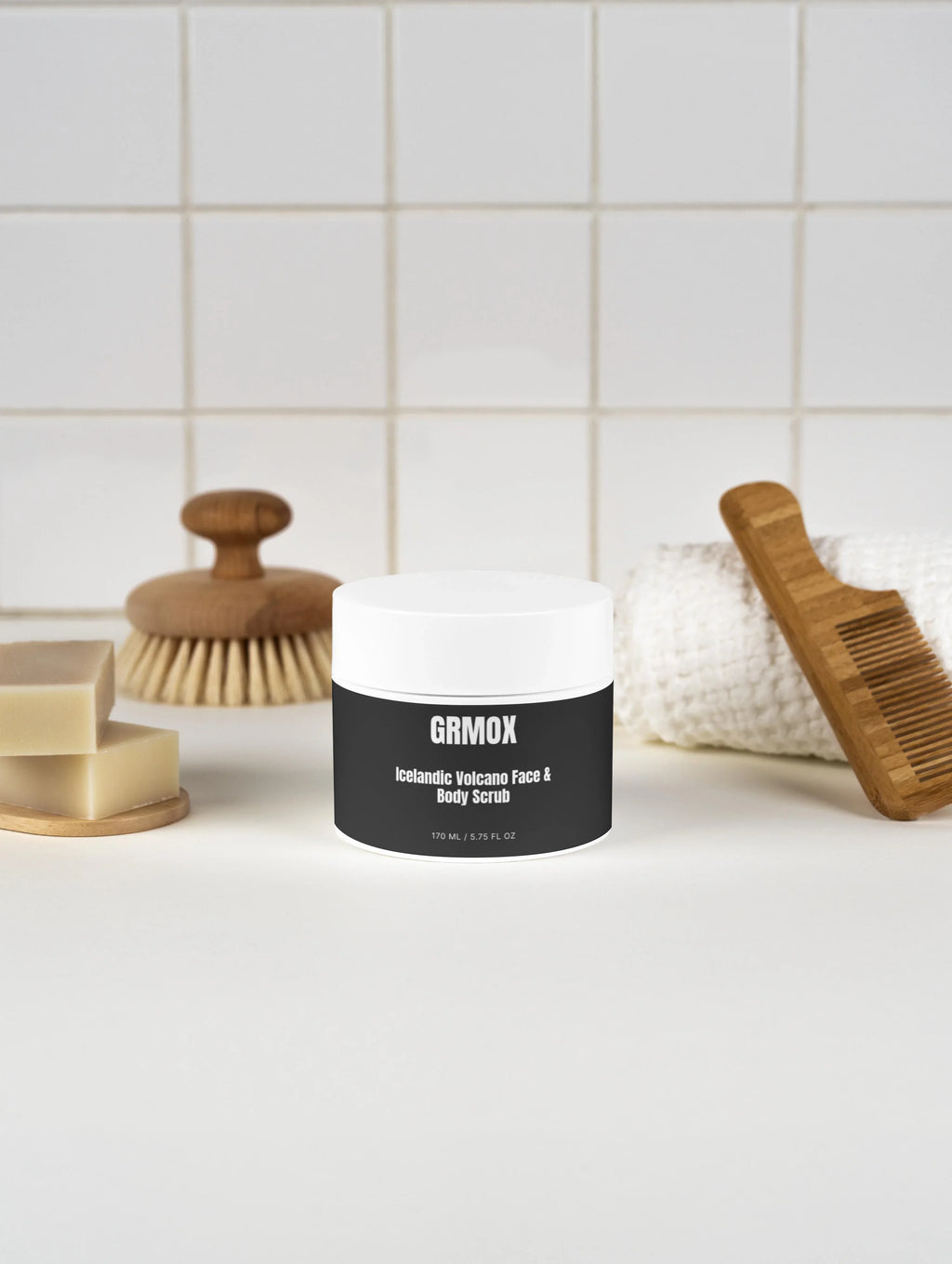 GRMOX Icelandic Volcano Face & Body Scrub in white jar with wooden brush, soap, and towel on tiled bathroom counter